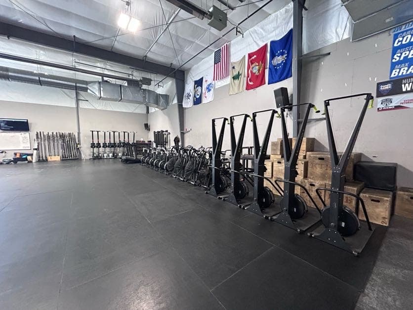 A long row of ski ergs, assault bikes, and rowers lined up along the wall under the service flags