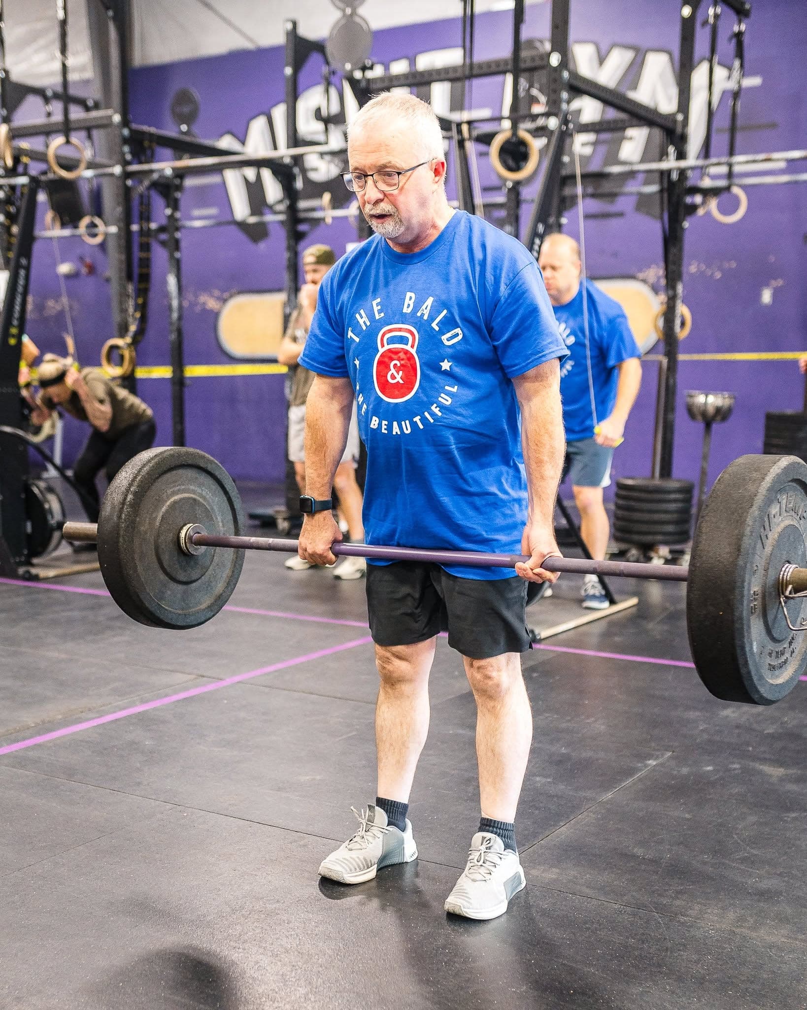 An older Misfit member in a Bald and Beautiful shirt standing over a loaded barbell on the floor, under the gym's purple mural