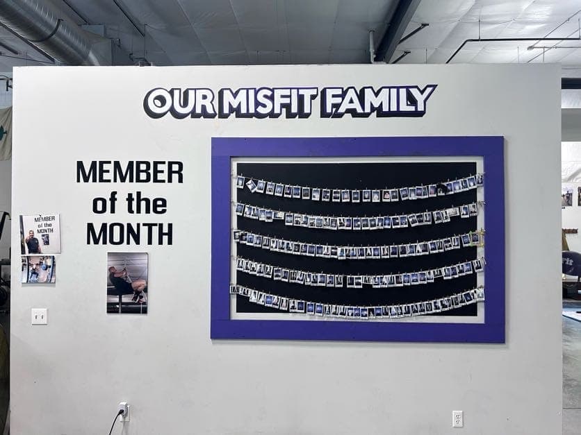 The Our Misfit Family wall: a purple-framed board strung with rows of Polaroid portraits of members, beside a Member of the Month display