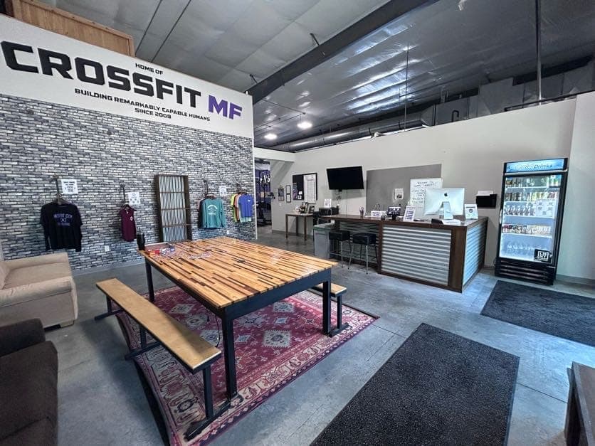 The lobby: the Home of CrossFit MF wall sign reading Building Remarkably Capable Humans Since 2009, a long communal table, the front desk, and the shirt rack and drinks cooler of the honor-system store