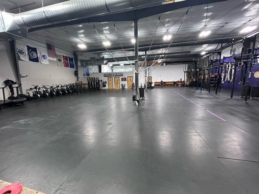 The same floor at standing height: a rig with gymnastic rings down the right side, the cardio row and the Strength in Numbers wall on the left, and a clear stretch of matted floor between them