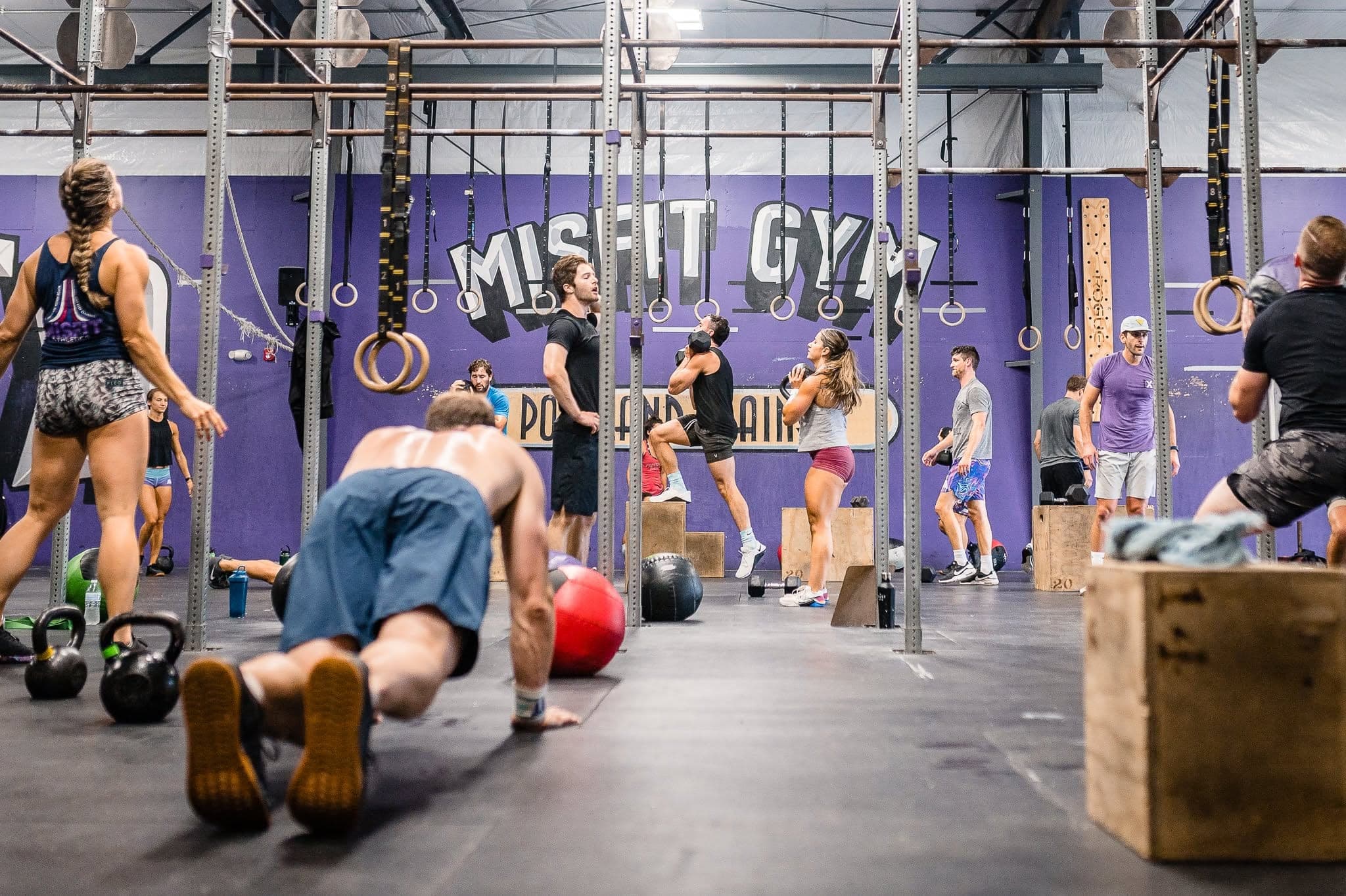 A full class working across the main floor under the Misfit Gym mural, one member mid push-up in the foreground while others work on the rigs and boxes behind