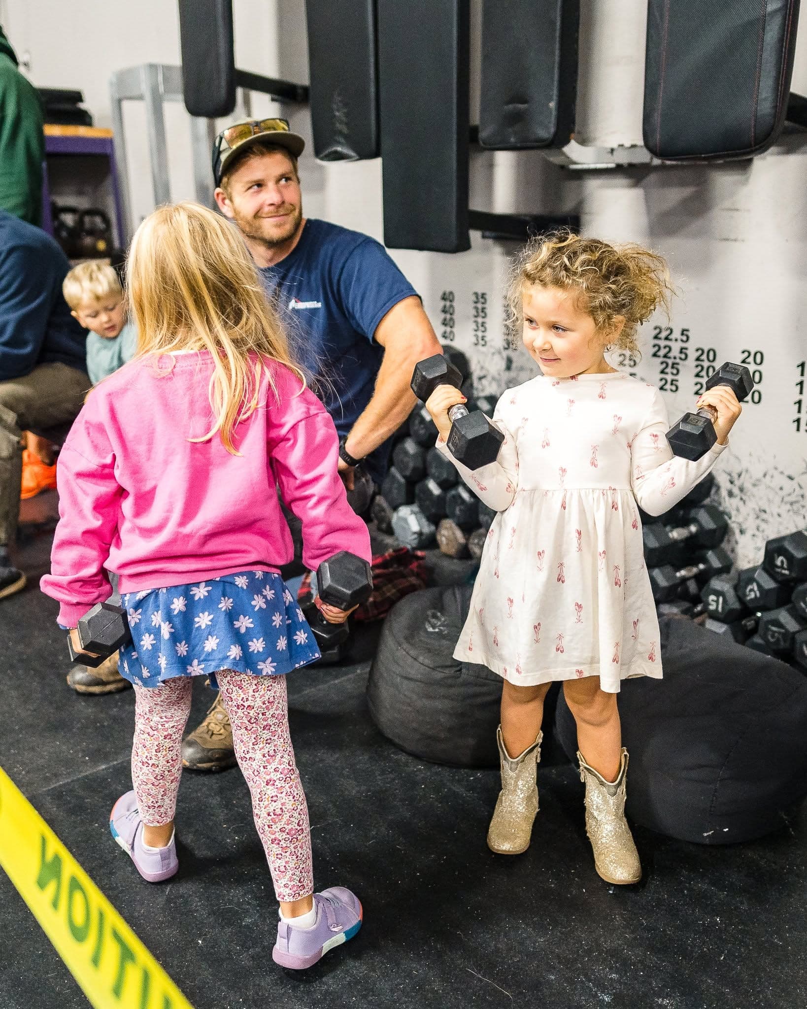 A coach in a cap smiling with three young children on the gym floor, two of them holding small dumbbells beside a rack of medicine balls