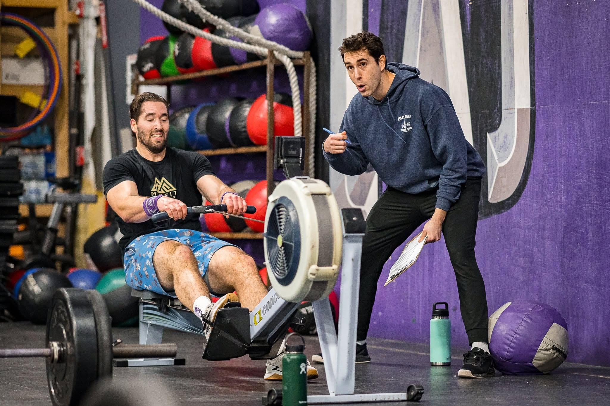 A coach with a clipboard leaning in to talk to one member mid-effort on the rower, the two of them working alone on the gym floor