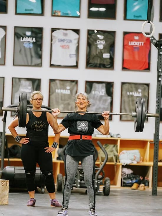 Misfit member pressing a loaded barbell overhead while a friend looks on